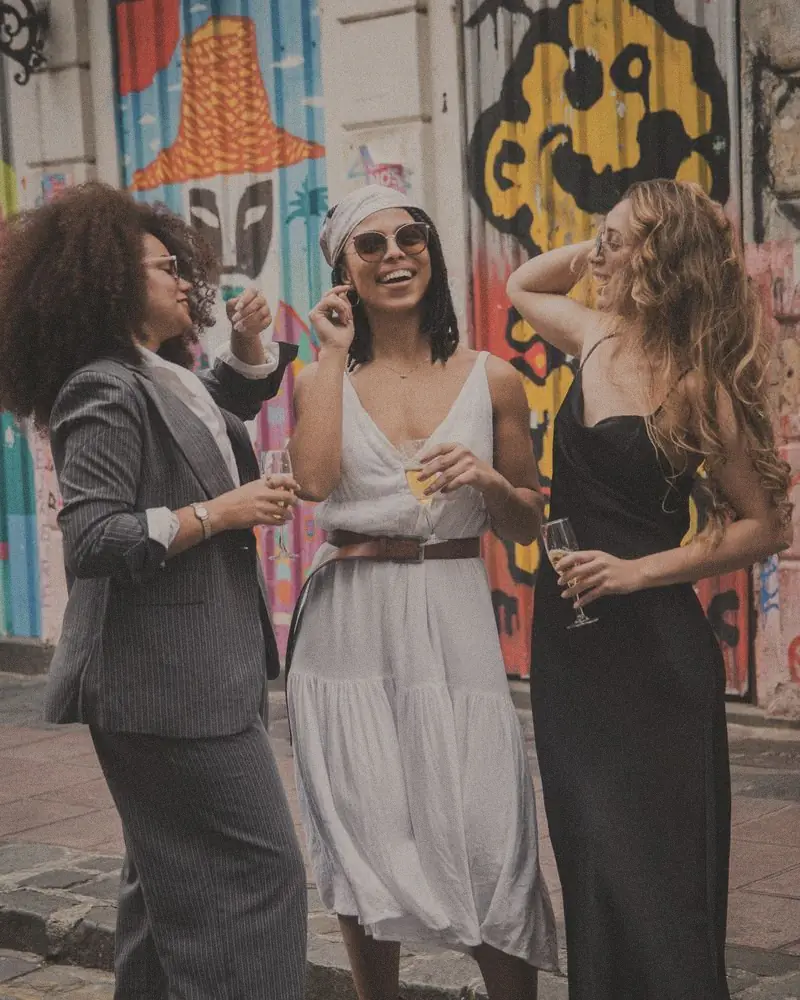 Trois amies qui rient une coupe à la main dans une rue pavée, la future mariée en blanc
